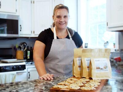 Woman in apron in kitchen showing fresh backed cookies next to bagged cookie kits