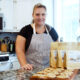 Woman in apron in kitchen showing fresh backed cookies next to bagged cookie kits